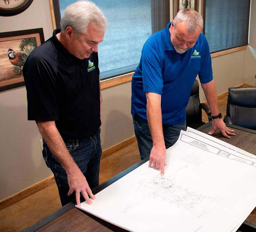 Two men in casual shirts discuss plans while pointing at a large architectural drawing on a conference table in a well-lit room.