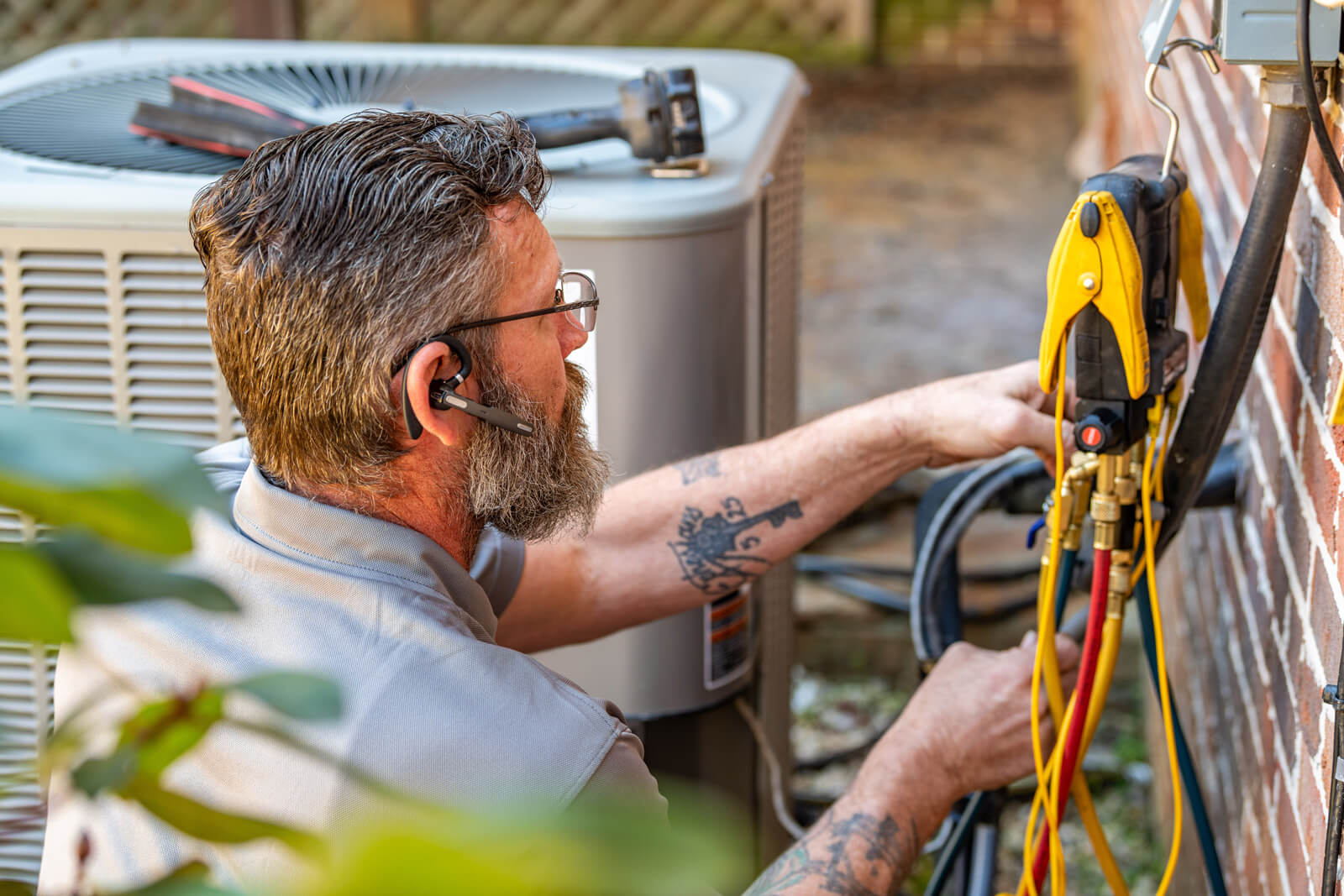 technician-fixing-hvac-outdoor-sideview A bearded man wearing glasses repairs an air conditioner, focusing on HVAC maintenance.