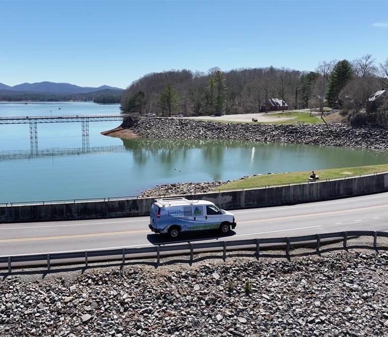 A van drives along a road next to a serene lake surrounded by mountains and trees under a clear blue sky.