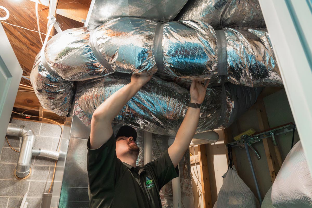 A man in gloves works on an air duct while servicing HVAC equipment in a residential setting.