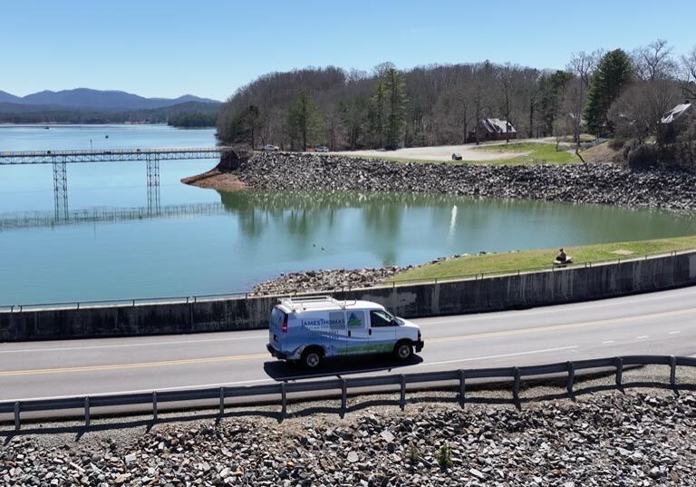 Morganton A van drives along a road next to a serene lake surrounded by mountains and trees under a clear blue sky.